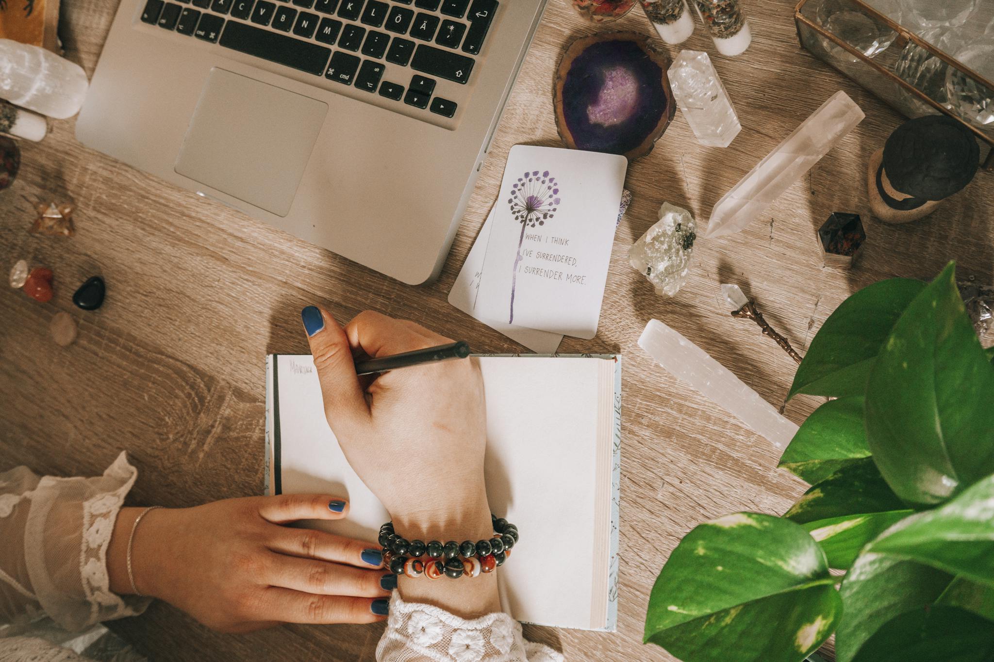 Woman's hand writing in a journal surrounded by crystals, plants, and laptop on a wooden desk.